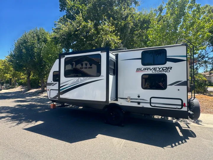 A white Surveyor Legend RV rental travel trailer parked on a street with trees under a blue sky.. Forest River Surveyor Legend 2023
