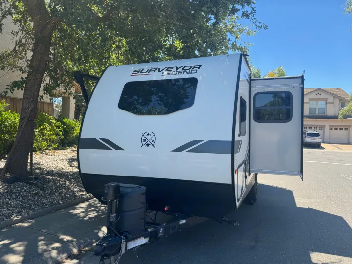 White Surveyor Legend travel trailer for RV rental, parked outdoors on asphalt, with its entry door open.. Forest River Surveyor Legend 2023