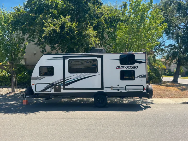 White "SURVEYOR LEGEND" travel trailer (rv rental) parked on a sunny residential street, surrounded by green trees.. Forest River Surveyor Legend 2023