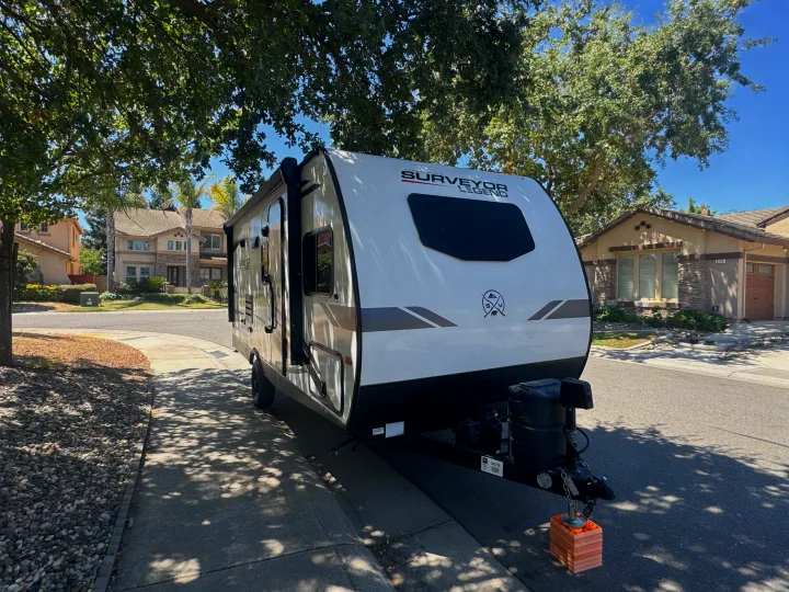 A white Surveyor Legend travel trailer, an RV rental, parked on a sunny suburban street shaded by trees.. Forest River Surveyor Legend 2023