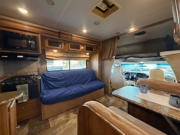 RV rental interior: well-lit kitchen with microwave & stove, blue sofa, dining table, and driver's cabin visible upfront.. Coachmen Leprechaun 2018