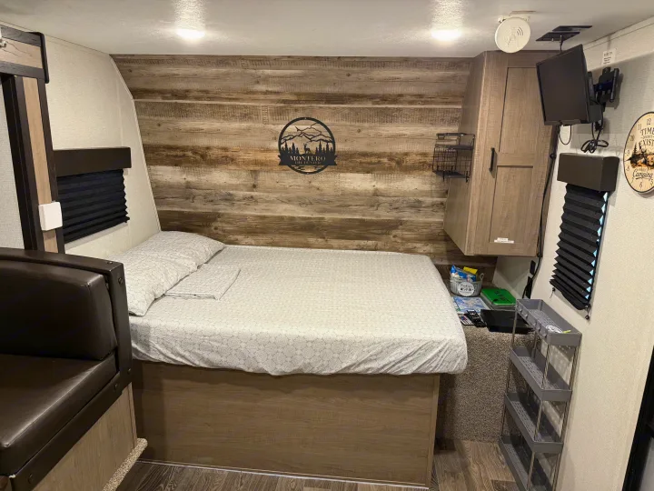 Inside this RV rental, a cozy bedroom with a patterned bed, rustic wood-paneled wall, TV, and ample storage for camping.. Keystone RV Hideout 2021