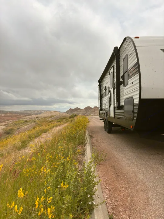 An RV rental rests on a road, offering a panoramic view of badlands landscape and yellow wildflowers under a cloudy sky.. Forest River Wildwood 2024