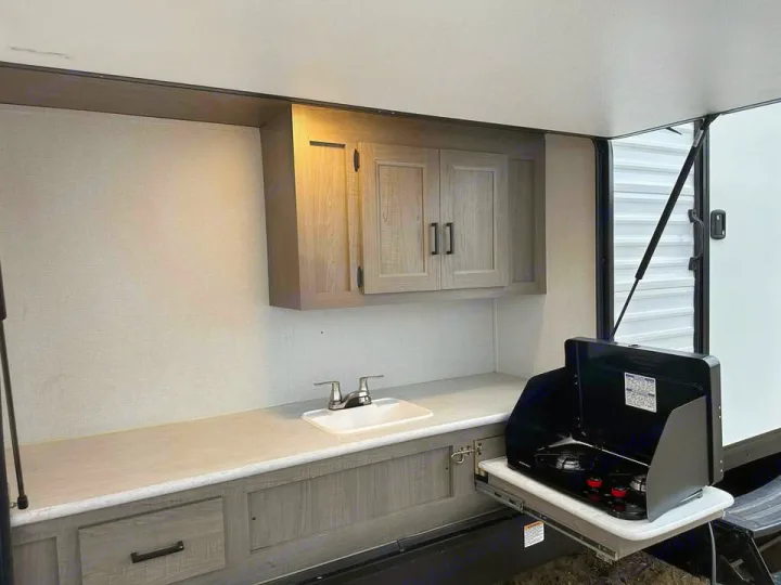 RV rental's outdoor kitchen area, featuring a countertop, sink with faucet, upper cabinets, and a pull-out two-burner stove.. Keystone RV Hideout 2021