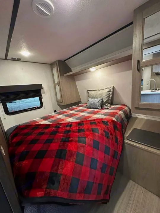 Charming RV rental bedroom with a cozy red plaid blanket, window, overhead storage, and mirrored closet.. Keystone RV Hideout 2021