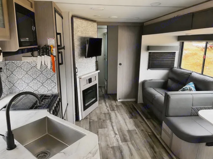 This modern RV rental interior features a kitchen with a patterned backsplash, sink, and stove, plus a gray sofa/dinette, fireplace, and wood floors.. Keystone RV 3055bs 2022