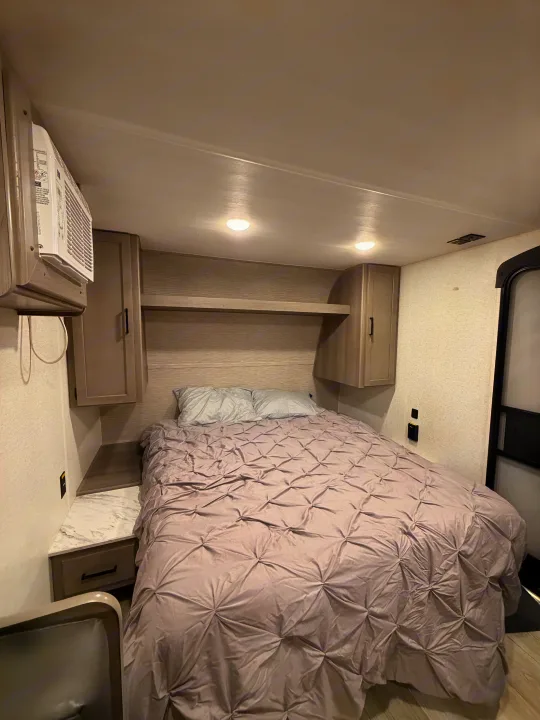 Cozy RV rental bedroom showing a bed with a grey comforter, overhead cabinets, a nightstand, and a wall-mounted AC unit.. Keystone RV Hideout Sport 2025