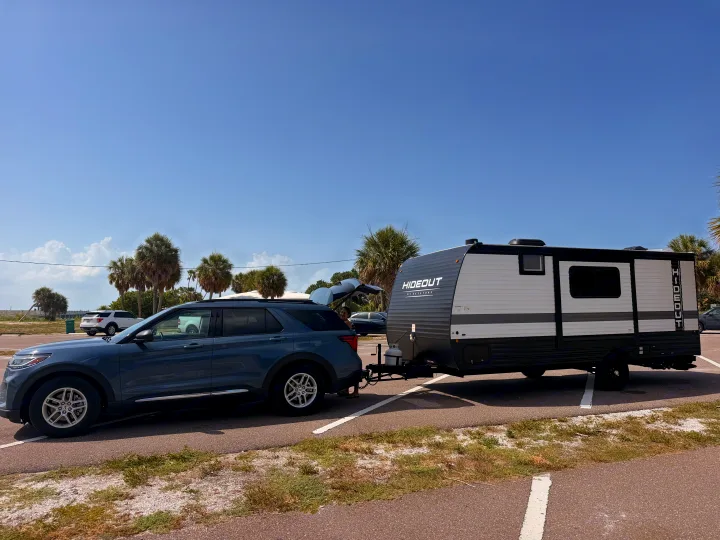 A blue SUV tows a 'Hideout' rv rental travel trailer in a sunny parking lot with palm trees, ready for an outdoor adventure.. Keystone RV Hideout Sport 2025