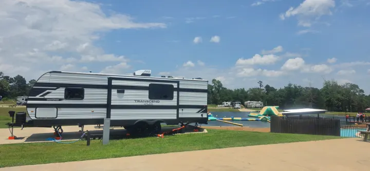 An RV rental at a scenic campsite by a lake with an inflatable water park, surrounded by trees and other RVs.. Grand Design Transcend Xplor 26BHX 2027