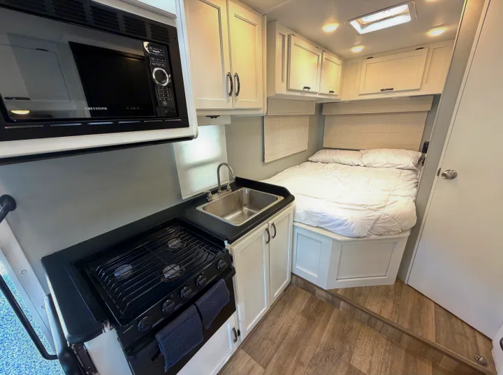 Modern rv rental interior featuring a compact kitchen with a microwave, stovetop, and sink, plus a cozy bed.. Thor Four Winds 2019