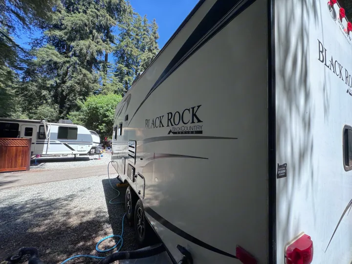 Black Rock Backcountry Series RV rental at a sunny campground. This travel trailer is nestled among tall trees with hookups.. Outdoors RV Blackrock 2017