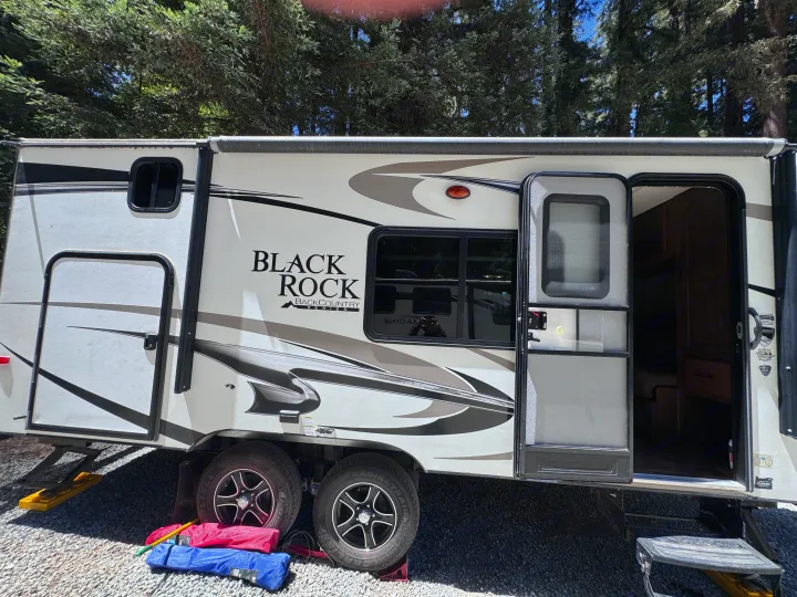 RV rental: A white Black Rock travel trailer with black/grey graphics, parked on gravel at a sunny campsite with trees, door open.. Outdoors RV Blackrock 2017