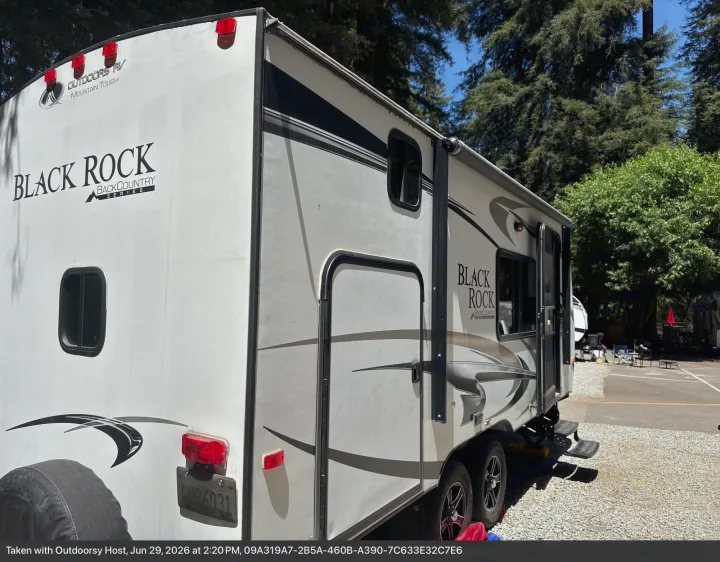 Back and side view of a white Black Rock Backcountry RV rental, parked at a sunny campsite amidst tall trees.. Outdoors RV Blackrock 2017