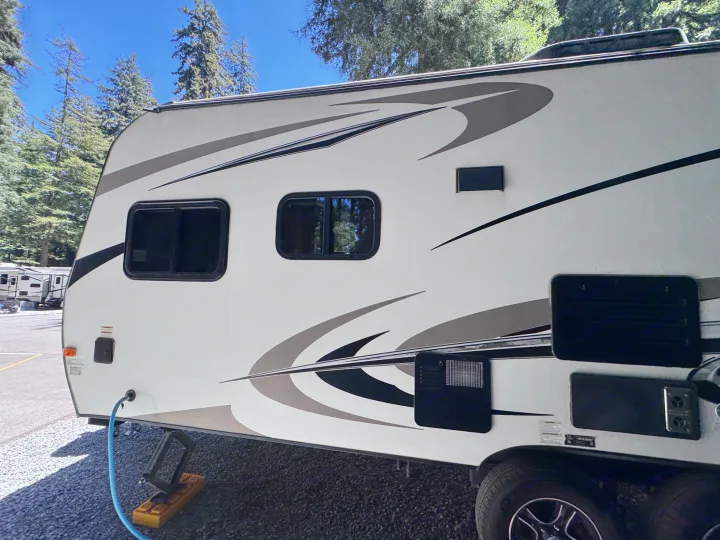 Side view of an RV rental, showing windows, vents, and a connected water hose at a scenic campground with tall pine trees.. Outdoors RV Blackrock 2017