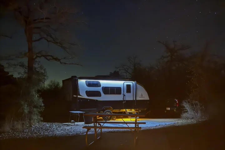 Starry night camping with an RV rental. Modern camper trailer with exterior lights on, illuminating the campsite picnic table.. Ember Overland 2023