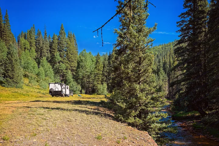 A white travel trailer (rv rental) parked in a sunny clearing, surrounded by dense green forest and a flowing river.. Ember Overland 2023