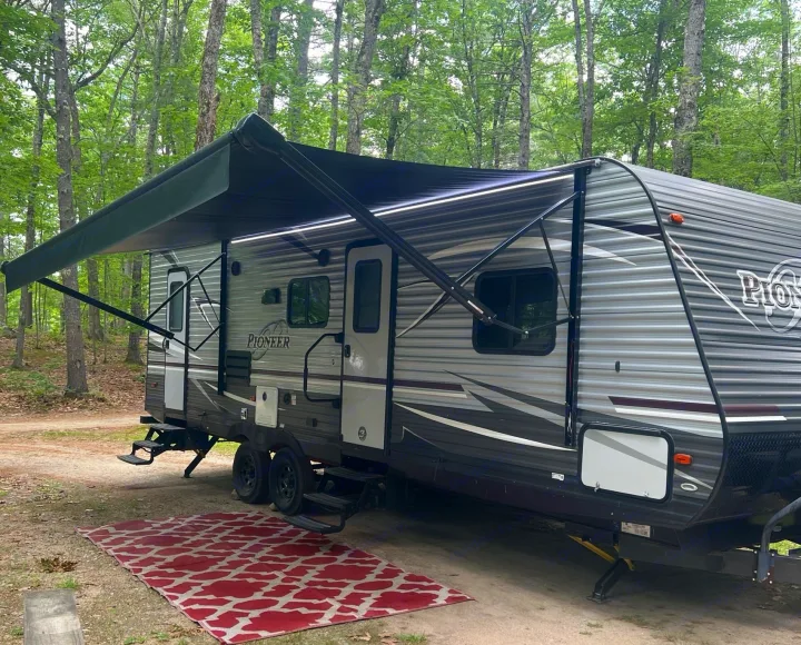 Discover this cozy rv rental travel trailer at a wooded campsite, featuring an extended awning with LED lights and an inviting outdoor rug.. Heartland RVs Pioneer 2018
