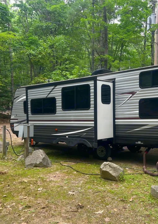 Grey travel trailer RV rental at a shady campground, hooked up to utilities. Lush trees surround the peaceful outdoor setting.. Heartland RVs Pioneer 2018