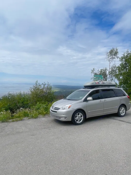 Silver minivan with a sticker-covered cargo box, ideal for an RV rental road trip, parked at a scenic overlook with a bay view.. Toyota Sienna Limited XLE Camper Van 2004