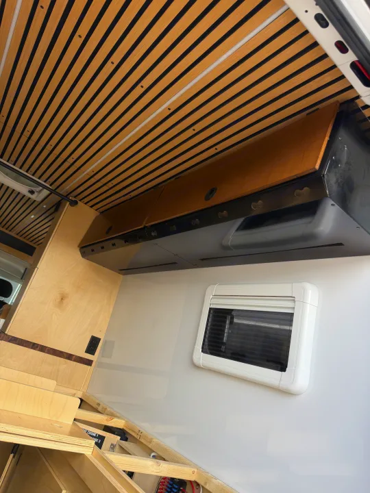 Modern RV rental interior showing a stylish slatted wood ceiling, custom overhead storage, and a white-framed window.. Safari Safari 2025