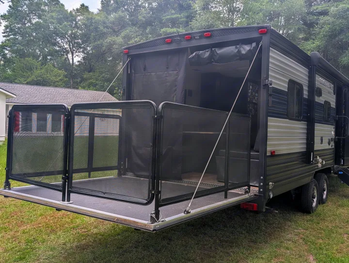 This grey and black rv rental toy hauler features its rear ramp lowered into a screened-in patio deck, parked on grass.. Forest River Cherokee Grey Wolf 2021
