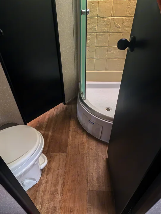 This RV rental bathroom features a white toilet, a compact rounded shower, and open black doors on a wood-look floor.. Forest River Cherokee Grey Wolf 2021
