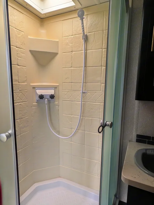 Compact RV rental shower interior featuring beige textured walls, corner shelf, handheld shower head, and water control panel.. Forest River Cherokee Grey Wolf 2021