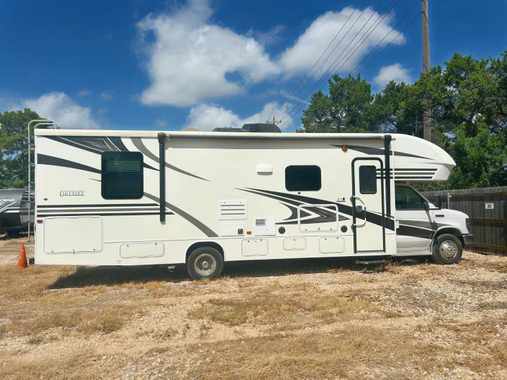 A white Class C RV rental, featuring black and grey graphics, parked on a dry, gravel lot under a bright blue sky.. Entegra Coach Odyssey 2019