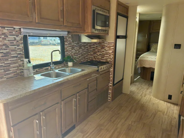 Explore this RV rental's well-appointed kitchen: double sink, stove, microwave, fridge, and backsplash, with a peek into the bedroom.. Entegra Coach Odyssey 2019