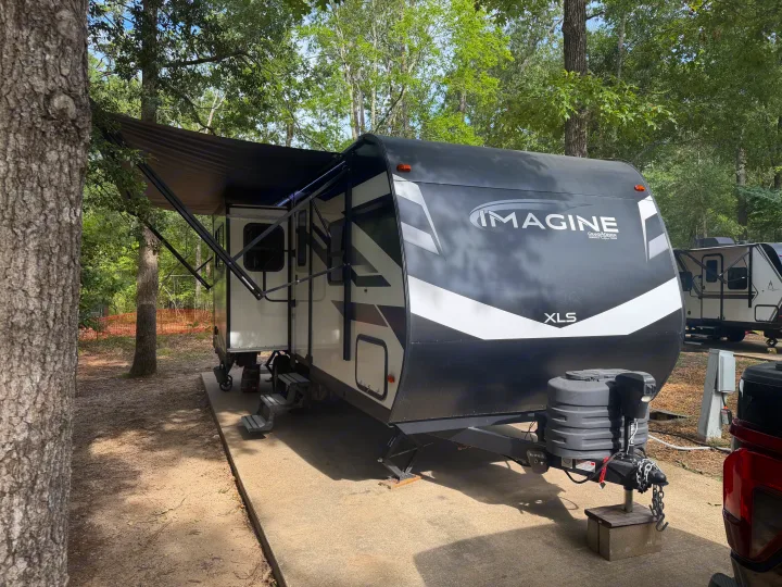 This Grand Design Imagine XLS travel trailer RV rental, with an extended awning, is parked on a concrete pad at a wooded campsite.. Grand Design Imagine XLS 2024