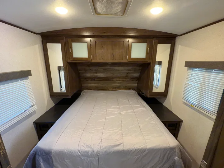 Inside a comfortable RV rental bedroom featuring a full bed, overhead storage cabinets, and side windows with blinds.. Keystone RV Passport Grand Touring 2020