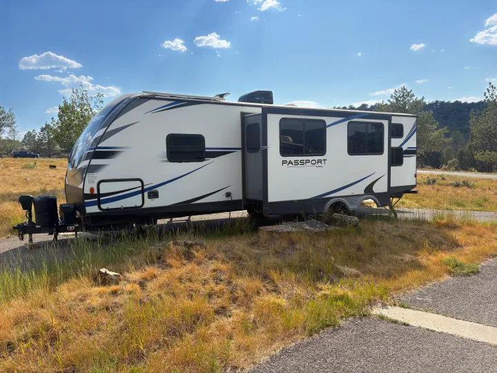 This white Passport travel trailer RV rental, extended with a slide-out, sits in dry grass under a blue sky for a relaxing getaway.. Keystone RV Passport Grand Touring 2020