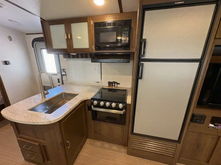 Inside this RV rental kitchen, you'll find a sink, stove, microwave, large fridge, and wood-look cabinets.. Keystone RV Passport Grand Touring 2020