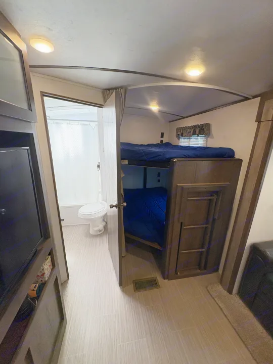 RV rental interior with bunk beds, blue bedding, a ladder, and a compact bathroom featuring a toilet and shower stall.. Keystone RV Passport Grand Touring 2020