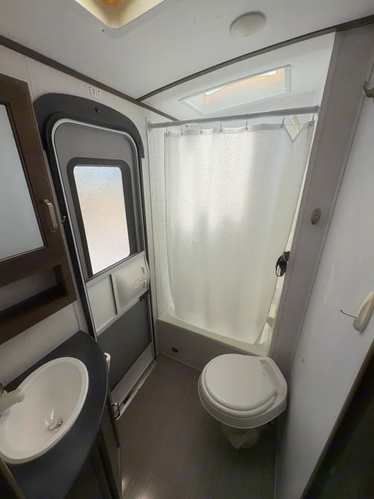 Inside an RV rental bathroom, showing a toilet, shower with curtain, and sink vanity. An 'EXIT' sign is above the door.. Keystone RV Passport Grand Touring 2020