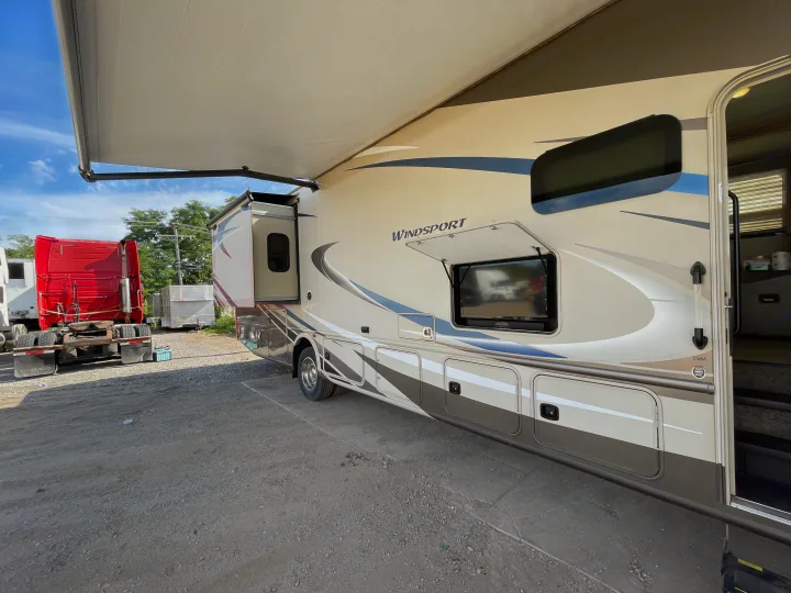 RV rental: A tan Windsport motorhome with an extended awning and slide-out is parked on a gravel lot, next to a red truck.. Thor Windsport 2019