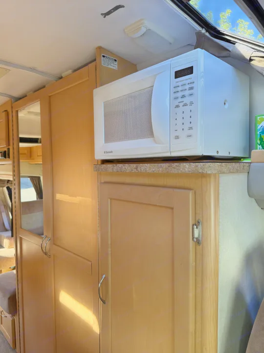 Compact Dometic microwave inside an rv rental kitchen, with wood cabinets and overhead light. Perfect for travel convenience.. Pleasure-Way Pleasure-Way Class B 2006