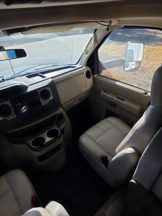 Interior of an RV rental: a bright cockpit with light seats, wood-trim dashboard, and clear views through the windshield.. Thor Chateau 2021