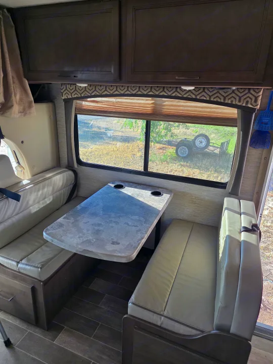 Cozy RV rental dinette with comfortable bench seating, a table with cup holders, and overhead storage, offering a scenic window view.. Thor Chateau 2021