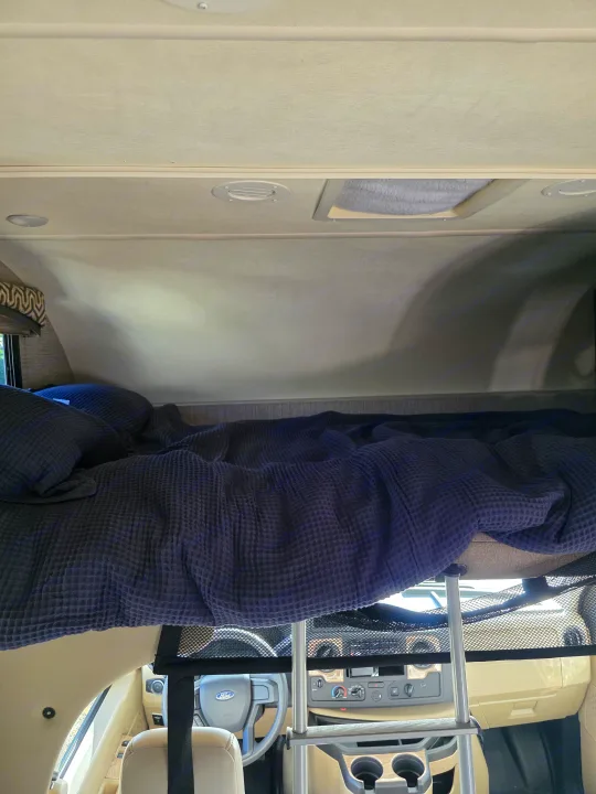 Cozy RV rental overhead bed setup with blue bedding, safety net, and visible front cabin dashboard.. Thor Chateau 2021