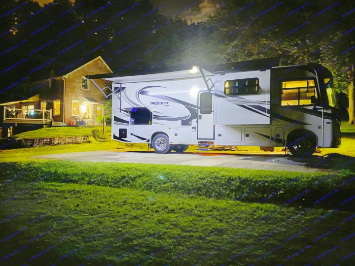 An illuminated RV rental parked at night, with an outdoor TV visible, next to a home on a grassy hill.. Jayco Precept 2022