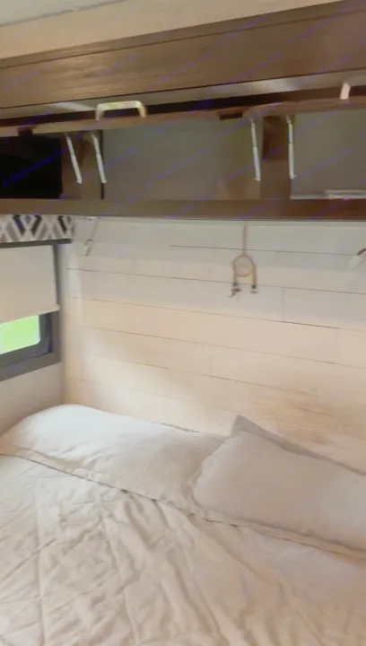 Cozy RV rental bedroom featuring a comfortable bed with light bedding, overhead storage cabinets, and a window with a green view.. Jayco Precept 2022