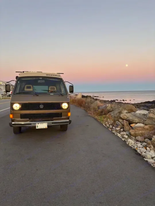 Experience freedom with this RV rental! A brown Westfalia van on the rocky Maine coast at sunset, with the moon above the ocean.. Volkswagen Westfalia 1985