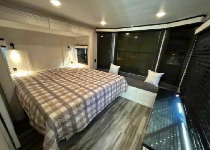 Spacious RV rental bedroom featuring a large plaid bed, window seat with pillows, windows, and wood-look flooring.. Jayco Jay Flight Bungalow 2025