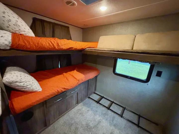 Experience comfort in this RV rental's cozy bunk beds, featuring vibrant orange bedding, a window with outdoor views, and a ladder.. Keystone RV Cougar 2018