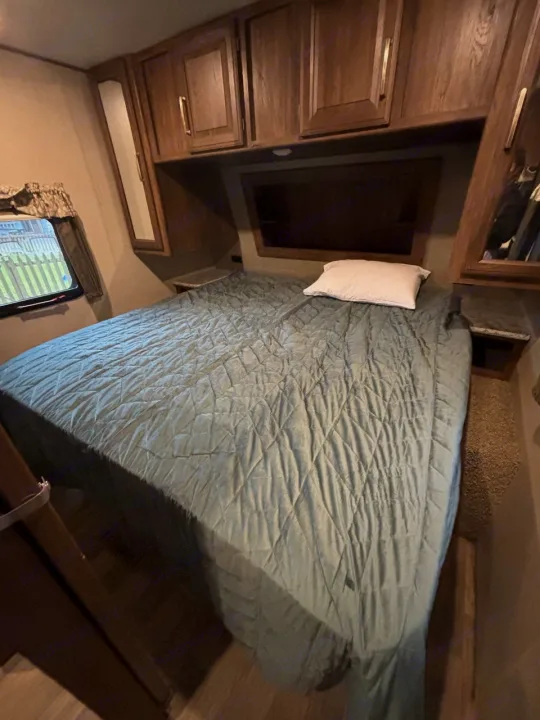 Cozy RV rental bedroom with a queen bed, quilted comforter, overhead wood cabinets, and a window showing outside.. Keystone RV Cougar 2018