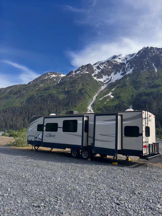 This RV rental, a travel trailer, is parked on a gravel site with majestic snow-capped mountains and dense green forests under a blue sky.. Keystone RV Cougar 2018