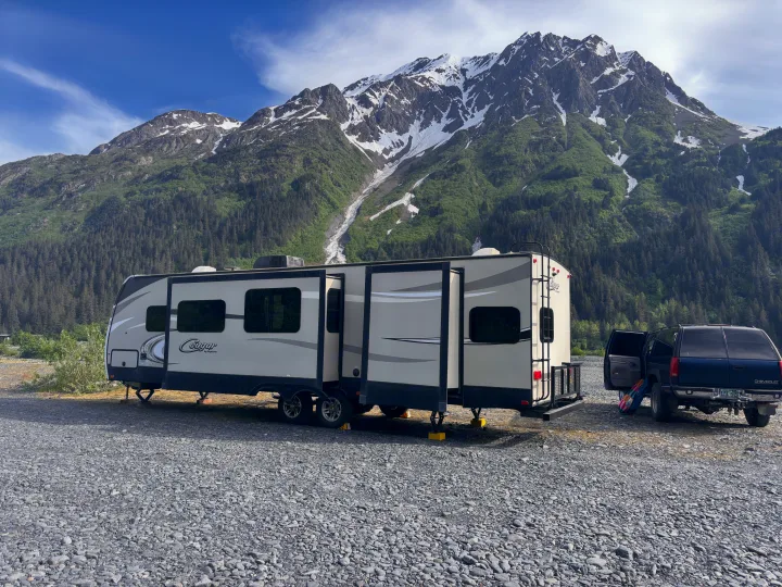 An RV rental and SUV are parked on a gravel lot before majestic, snow-capped mountains and green forests, under a blue sky.. Keystone RV Cougar 2018