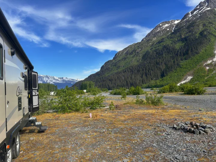 An RV rental is parked at a gravel campsite, offering stunning views of tall, tree-covered mountains and distant snowy peaks.. Keystone RV Cougar 2018