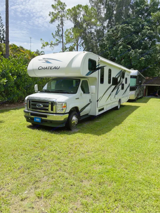 White Chateau RV rental parked on green grass, with another RV in the background, surrounded by trees under a bright, cloudy sky.. Thor Chateau 2025